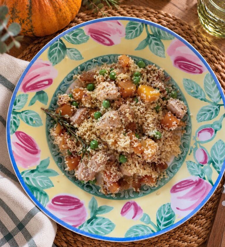Warm pumpkin salad with couscous, chicken, green peas, raisins and rosemary, served on a colourful floral plate.